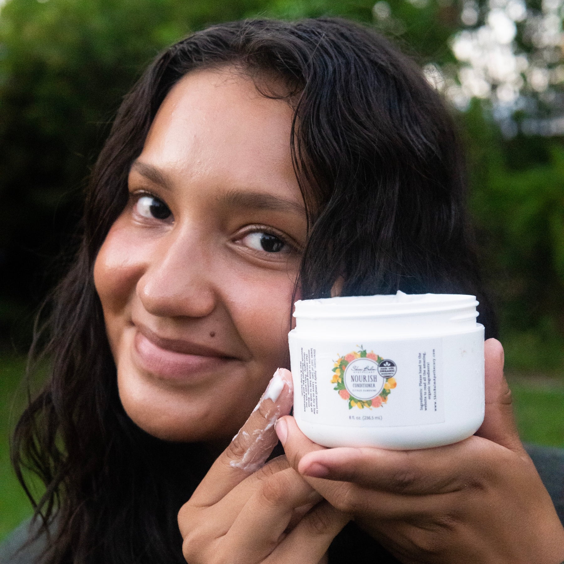 A close-up shot of a smiling woman holding a container of Citrus Sunshine Nourish Conditioner.